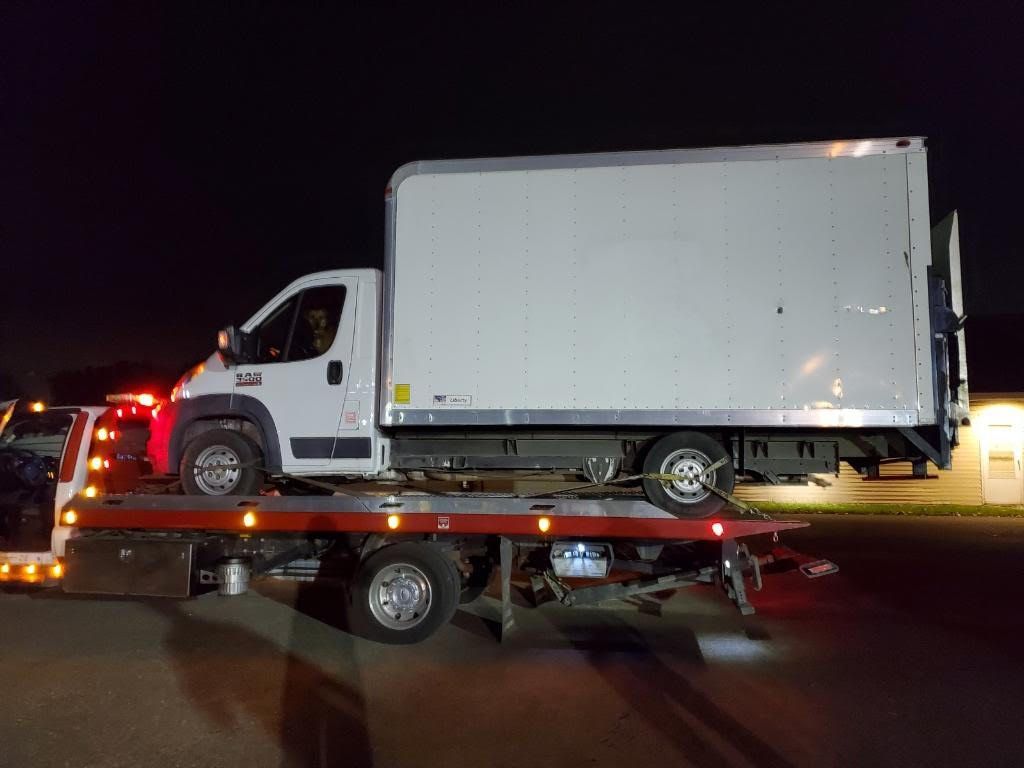 White box truck loaded on a flatbed tow truck at night with flashing red lights