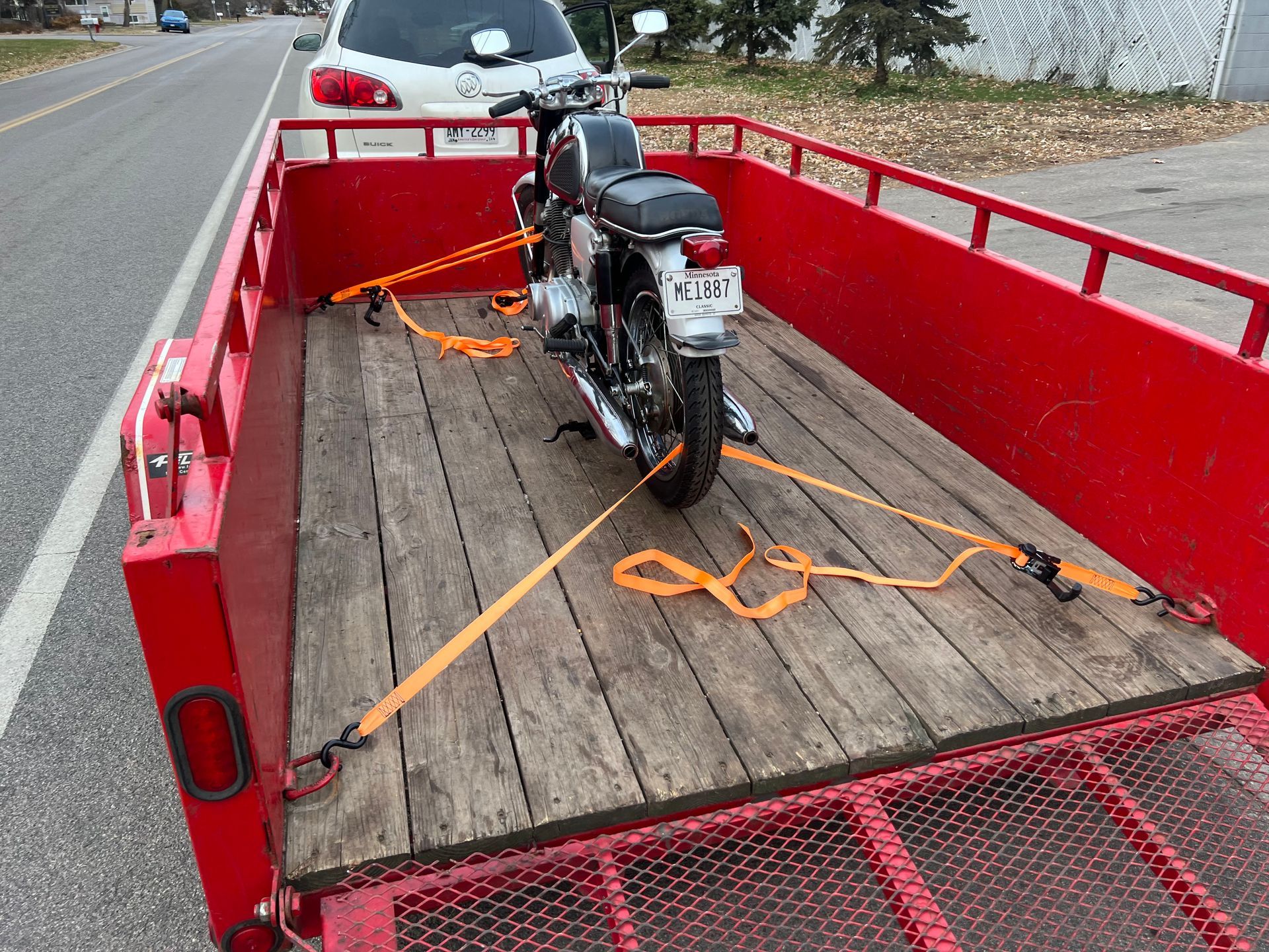 Motorcycle strapped on a red flatbed trailer with orange tie-downs beside a road