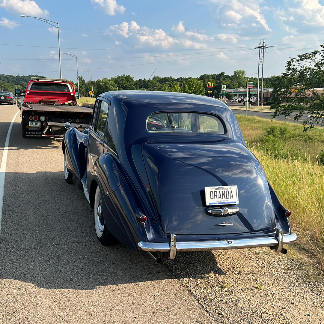 Blue vintage car being towed on a roadside, with a red truck ahead under a sunny sky.