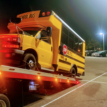 Yellow “school bus” tow truck on a flatbed at night in a parking lot, red lights glowing