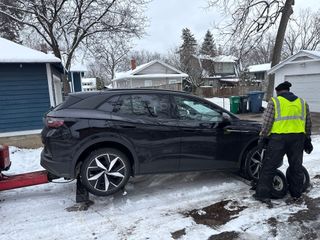 Person in a reflective vest works beside a black SUV stuck in snow in a residential driveway.