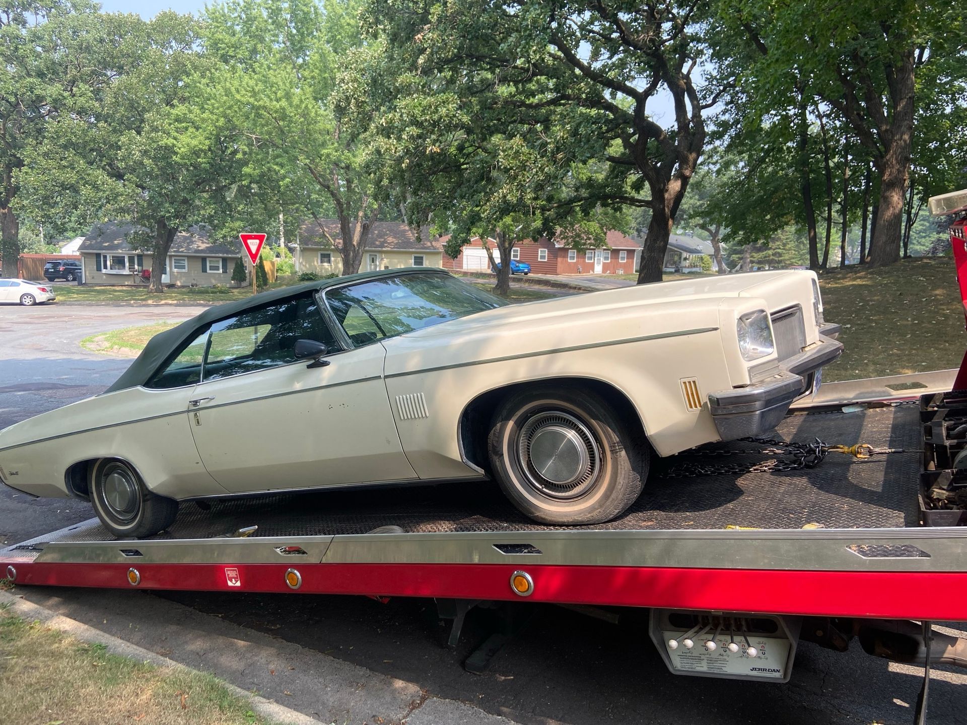 Vintage beige convertible on a red tow trailer in a tree-lined parking area.