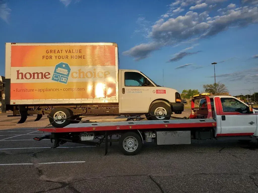 Tow truck carrying a beige box truck on a flatbed under a blue sky