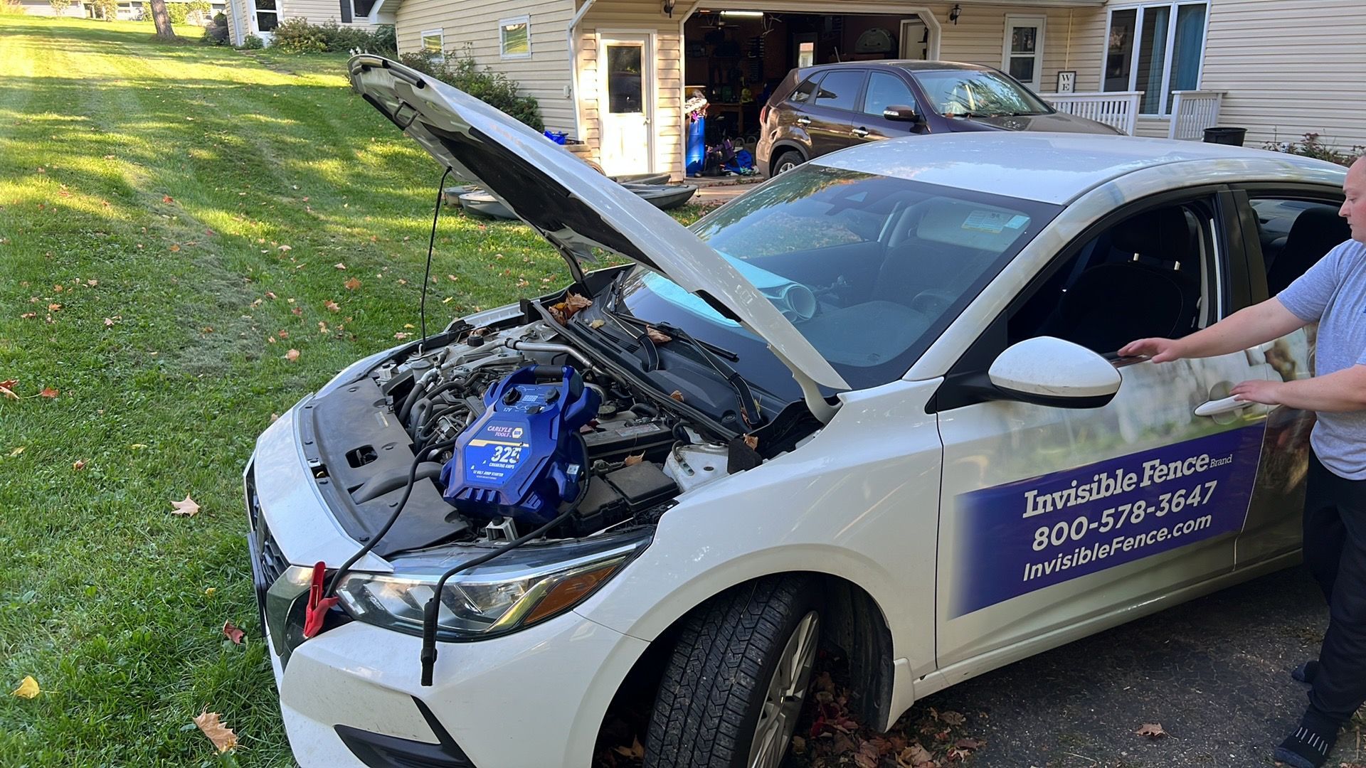 White car with open hood and front-end damage, displaying a roadside assistance sign in a driveway.
