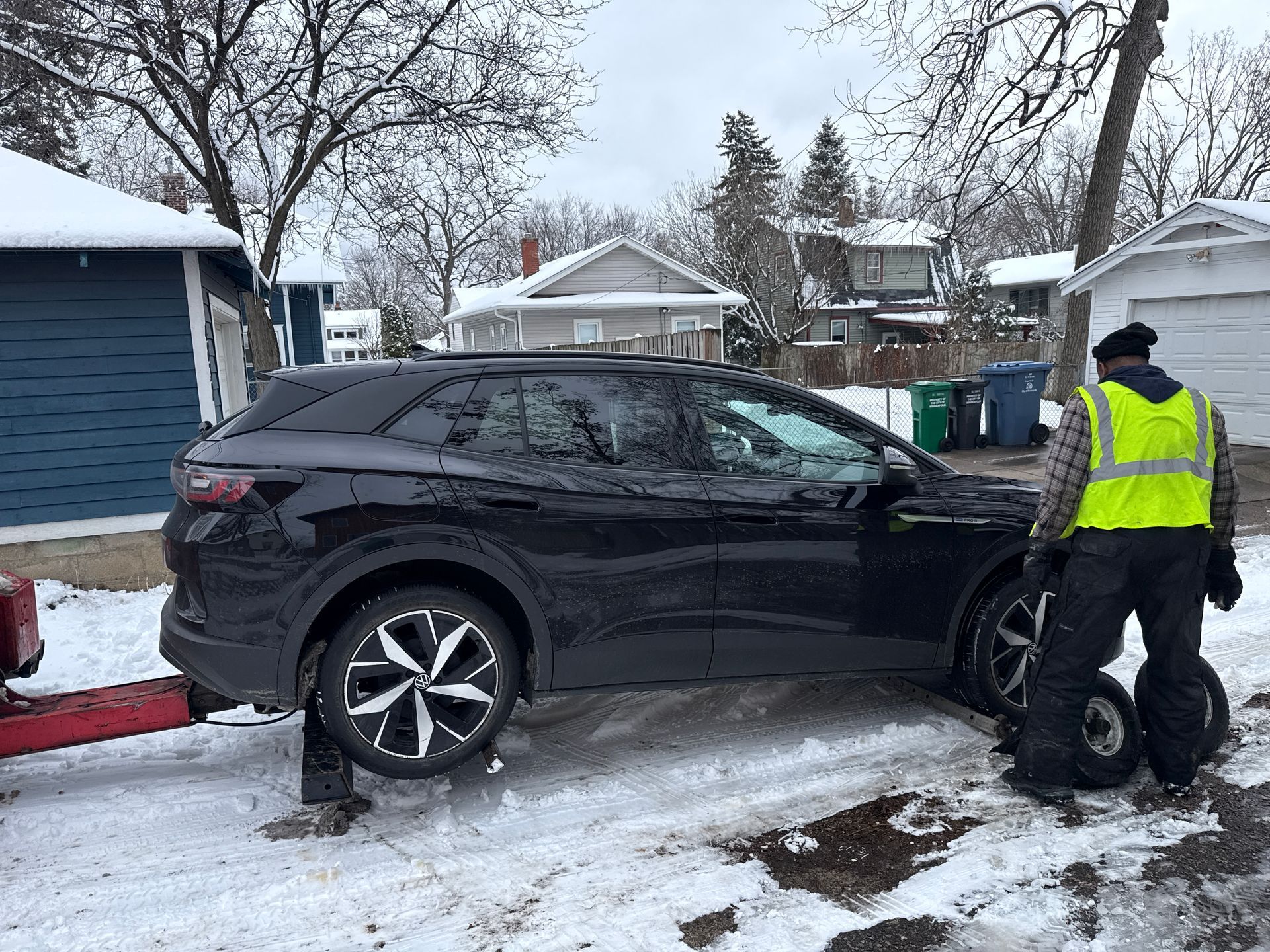Black SUV stuck in snowy driveway while a worker in a neon vest stands beside it