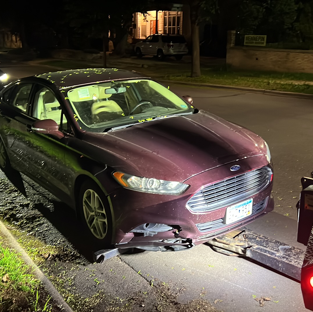 Maroon sedan with front bumper damage at night, parked by a curb under streetlights