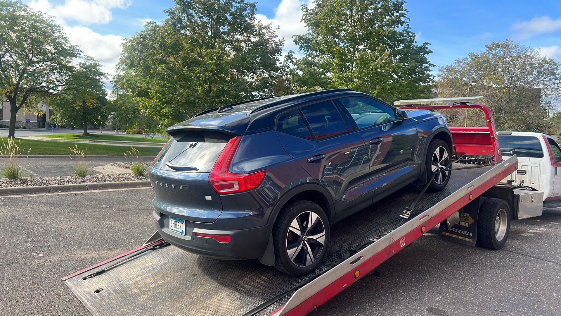 Dark blue SUV on a red flatbed tow truck in a parking lot with trees and houses behind it