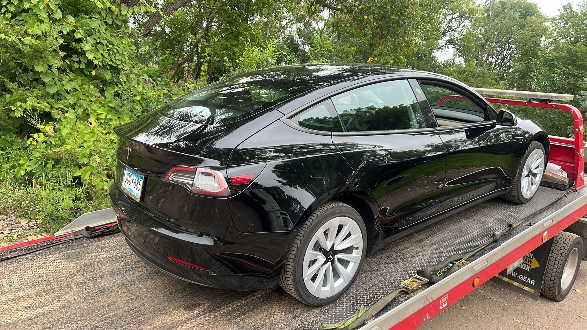 Black Tesla sedan strapped on a flatbed trailer beside a wooded roadside.
