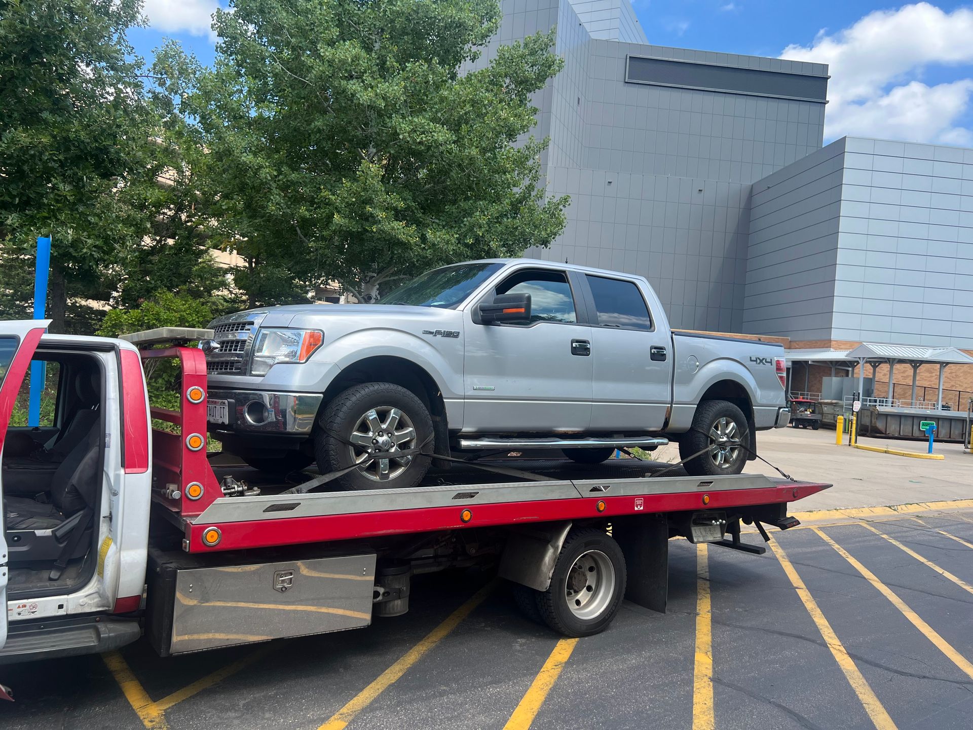 Silver pickup truck on a red tow flatbed in a parking lot beside a large building.