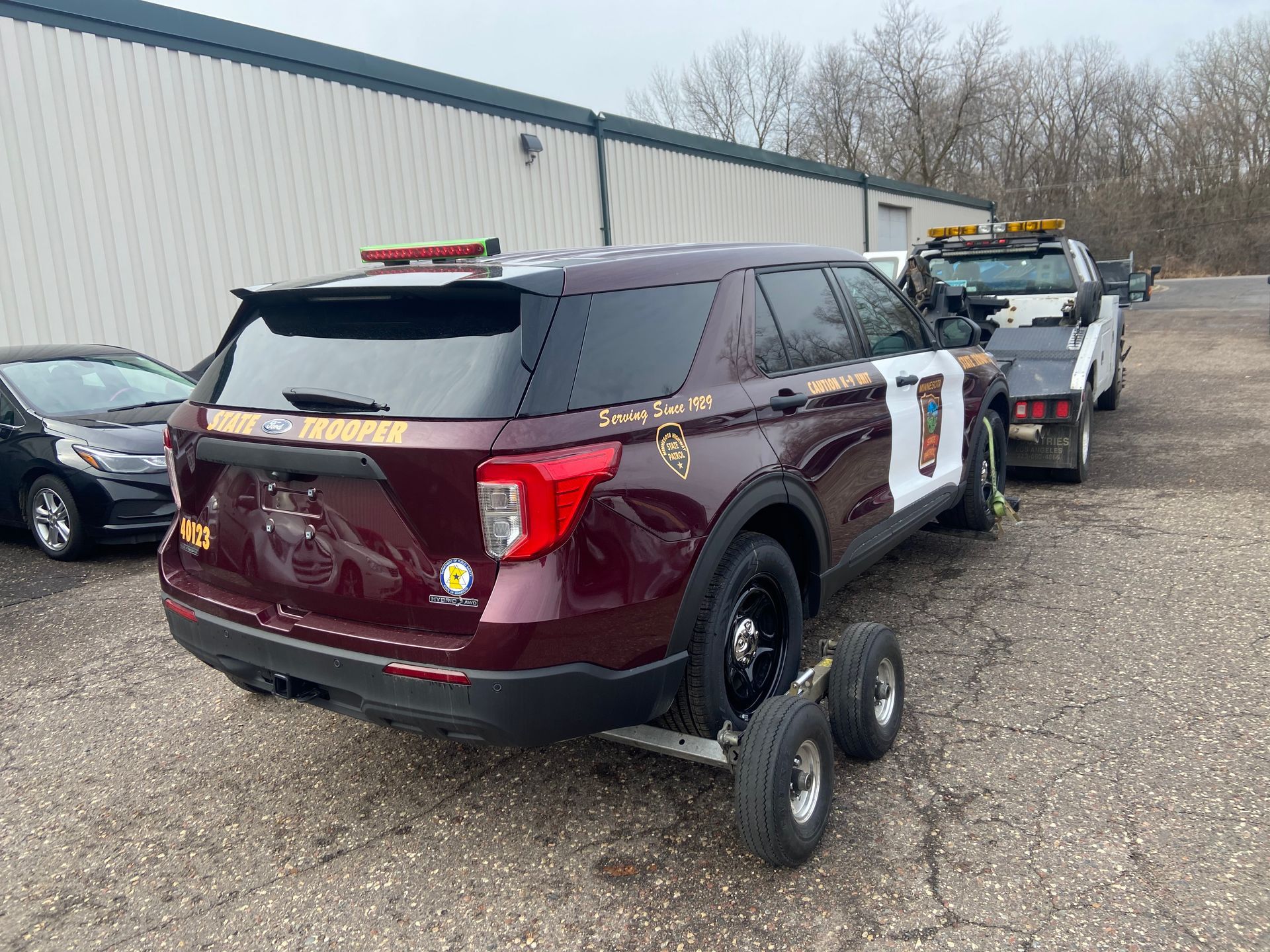 Maroon SUV on a dolly in a gravel lot, with another vehicle towing it near a warehouse.