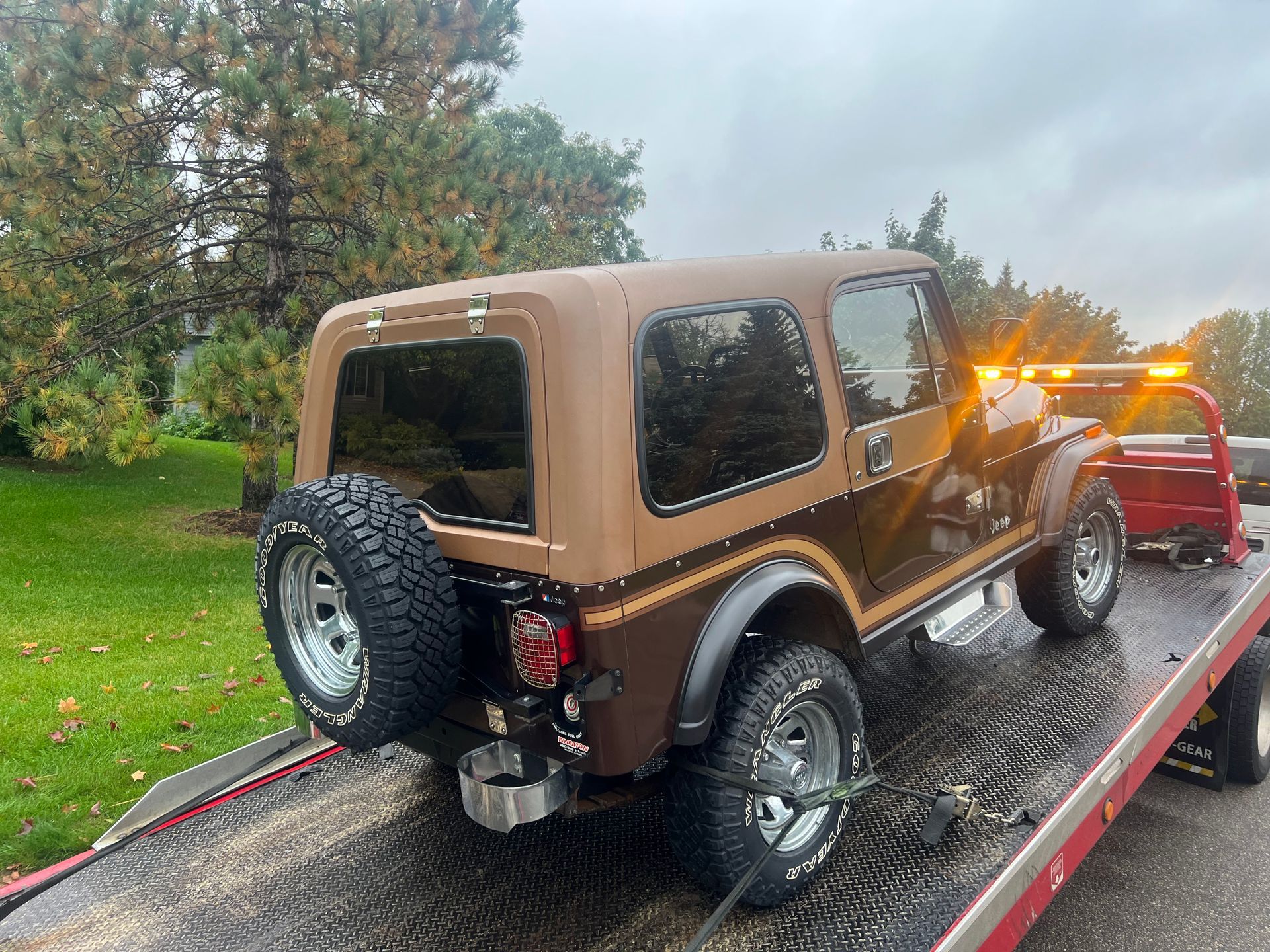 Brown Jeep Wrangler on a flatbed tow truck on a rainy roadside.