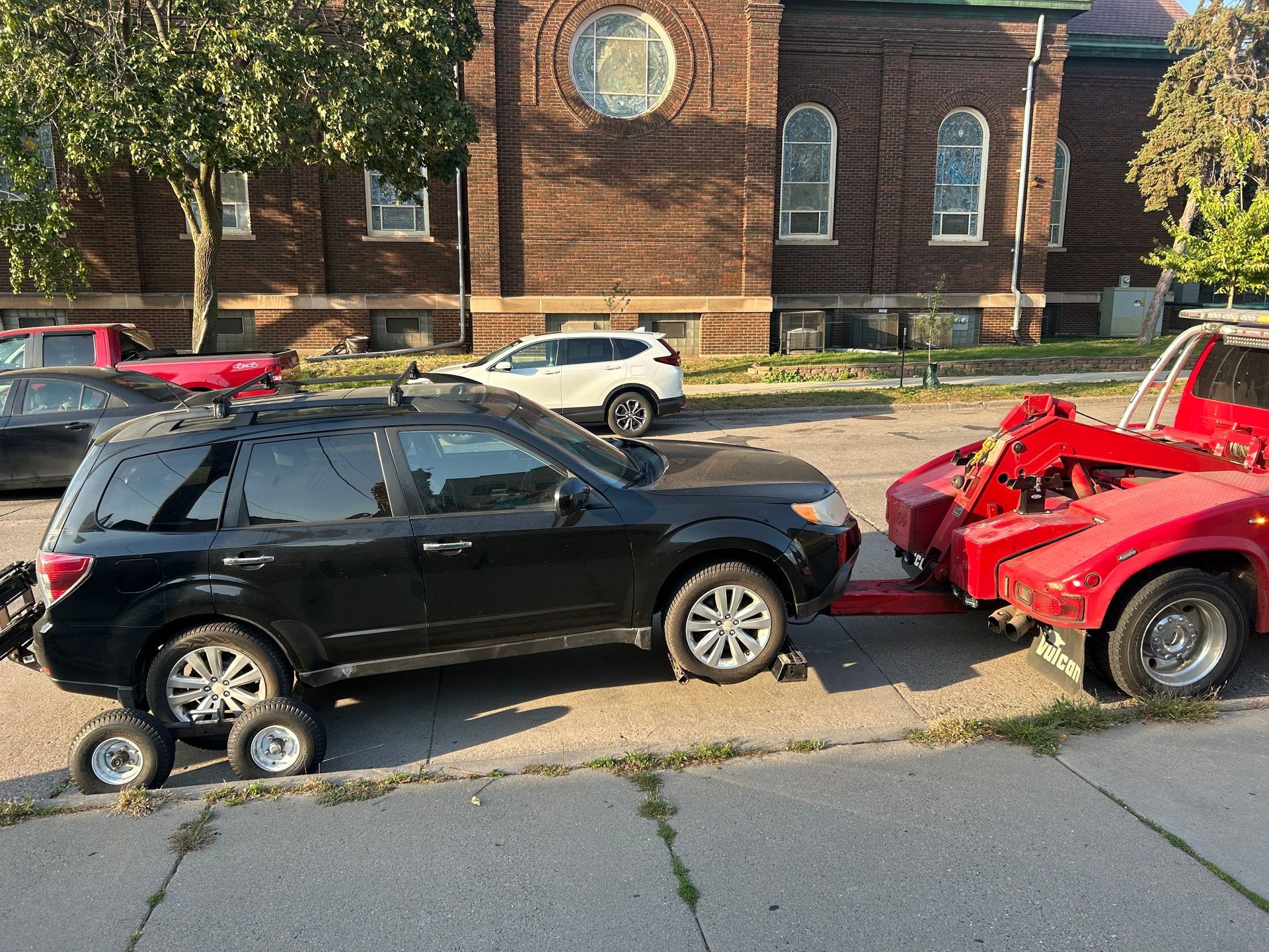 Black SUV being towed by a red tow truck on a residential street, with a brick building in the background