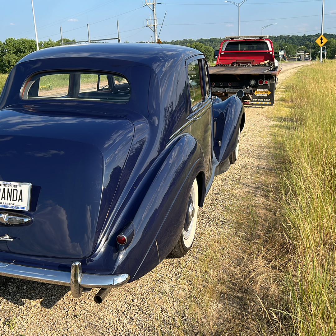 Blue vintage car towing a red truck on a roadside beside tall grass under a clear sky