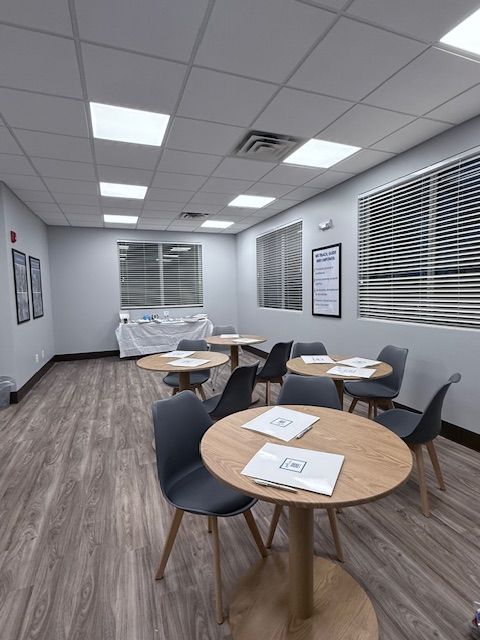 A well-lit waiting room with round tables, chairs, and window blinds; the floor is wood-like.
