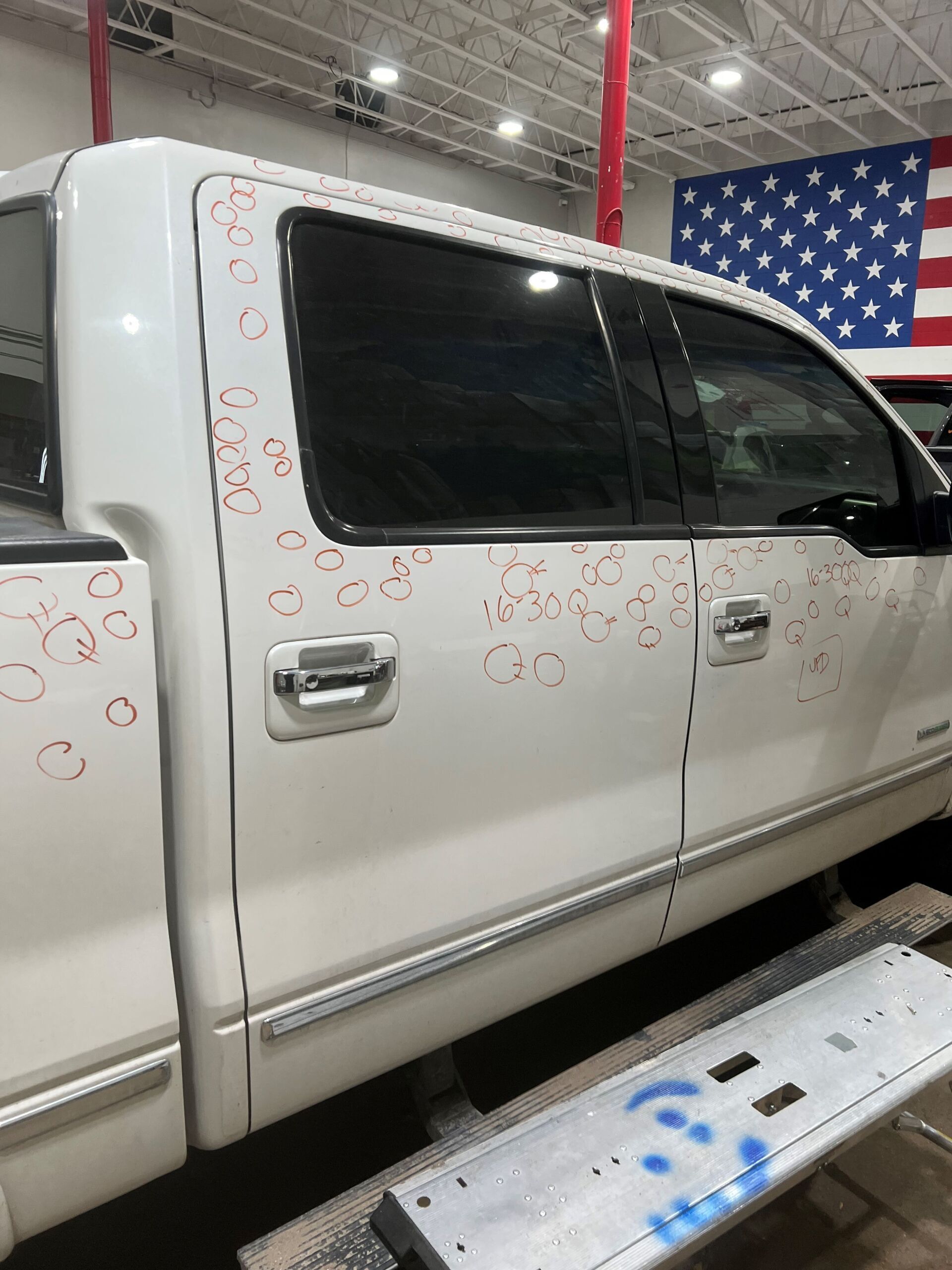 White pickup truck with circular decals, viewed indoors beside a metal step platform and an American flag backdrop