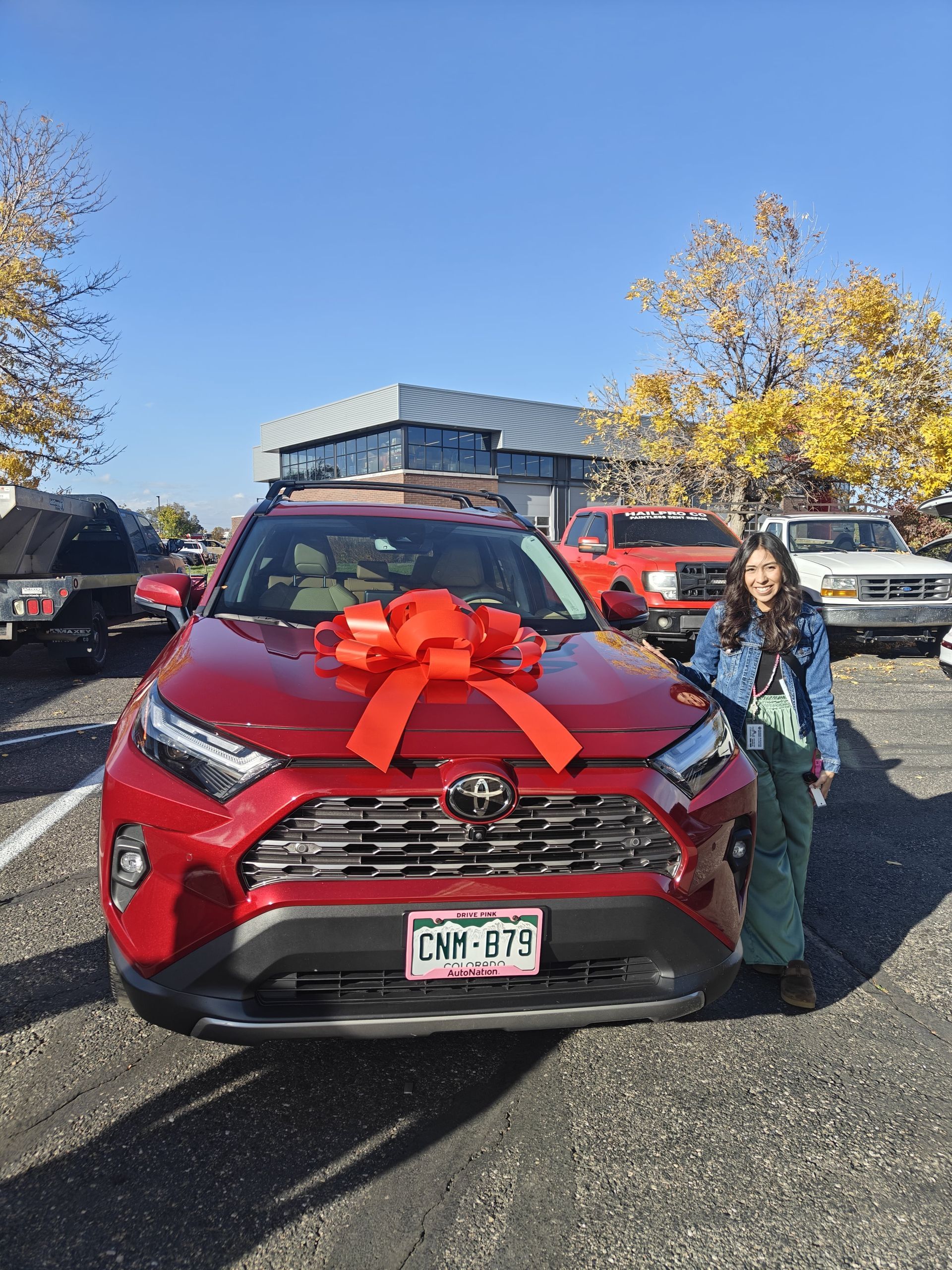 Red SUV with a large red bow; a person stands beside it in a parking lot on a sunny day.