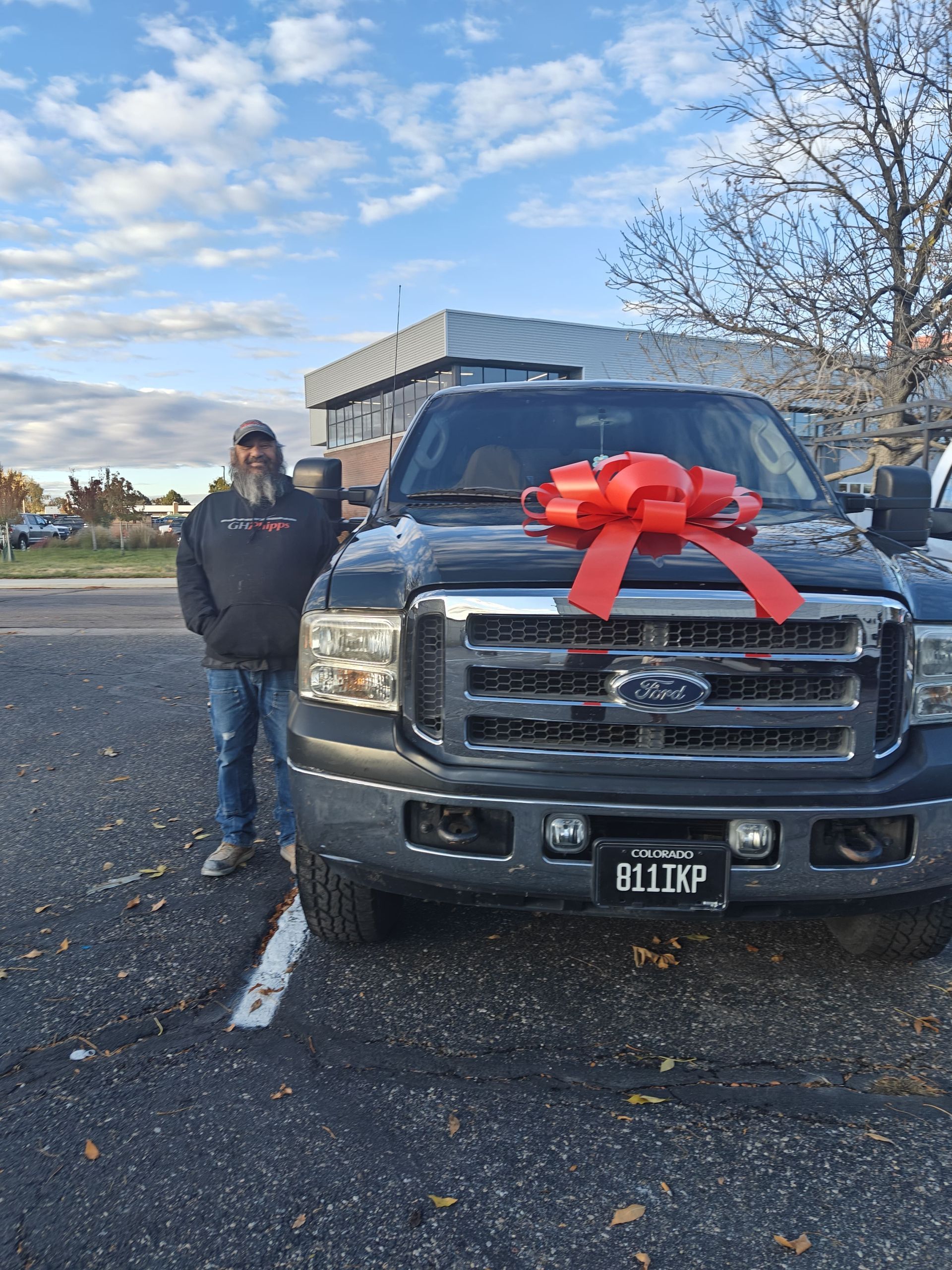 Person standing beside a black truck decorated with a large red bow in a parking lot.