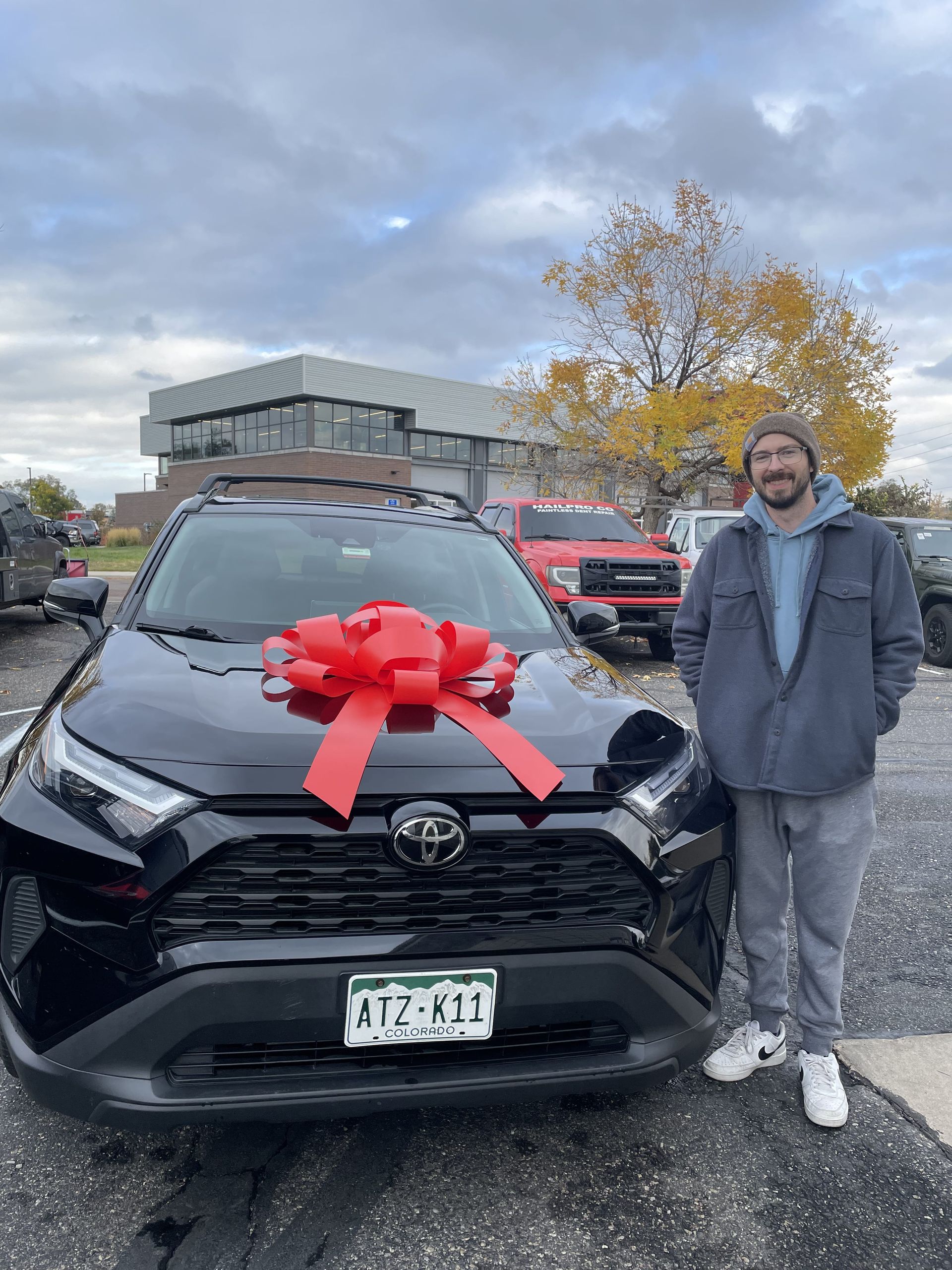 Man standing beside black Toyota with a red bow in a parking lot under a cloudy sky