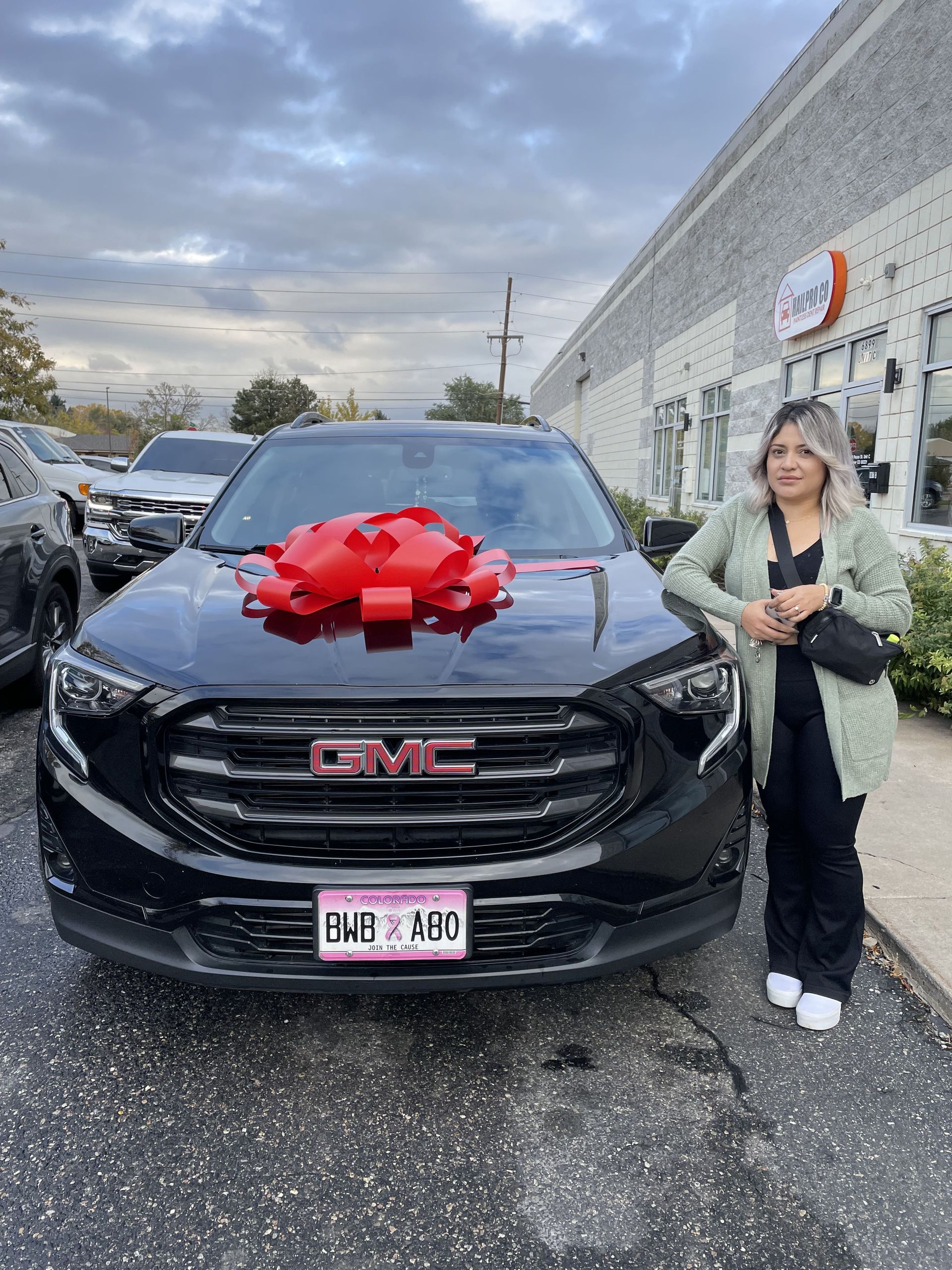 Woman stands beside black GMC SUV with a large red bow in a parking lot.