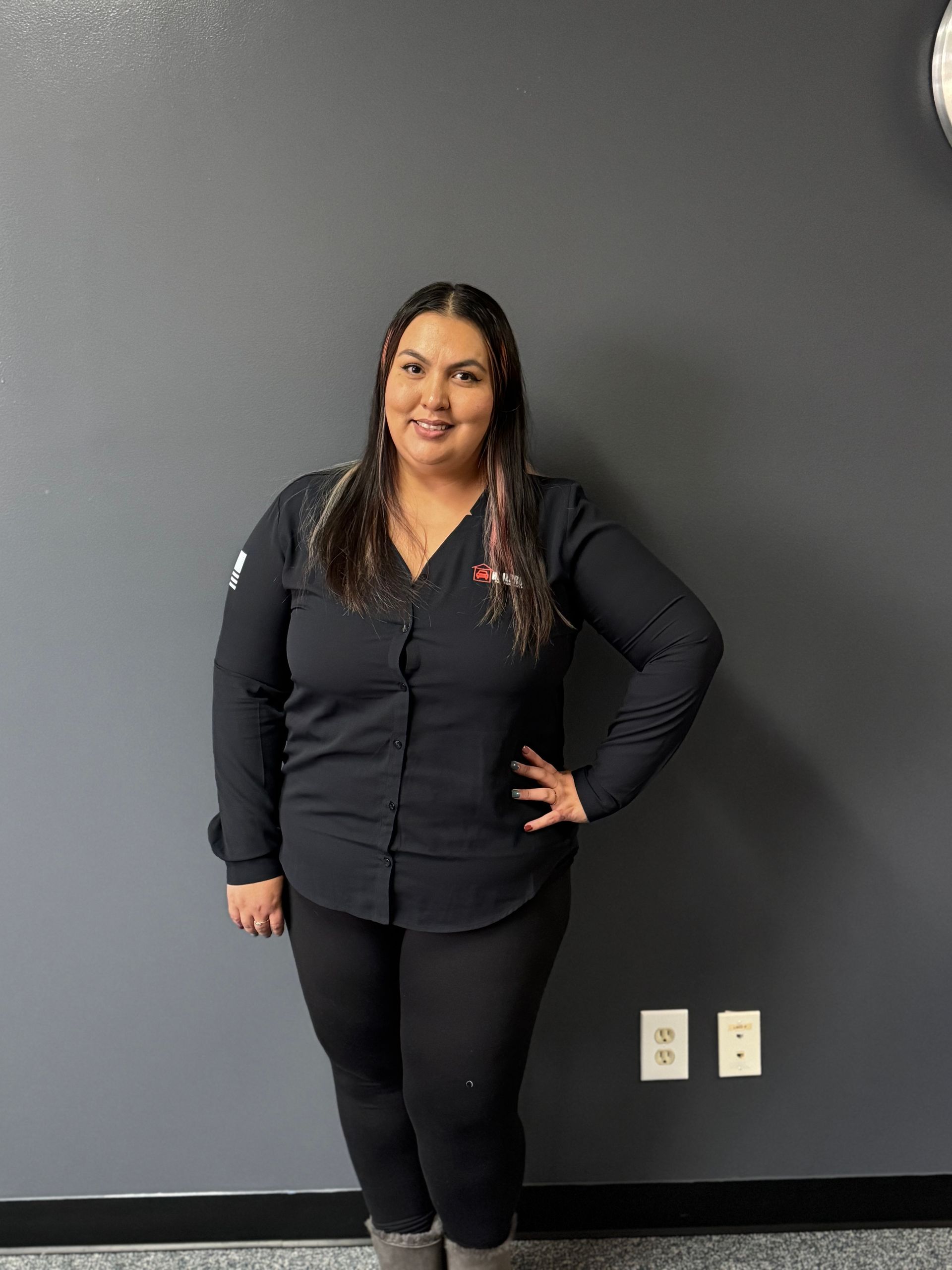Person standing against a gray wall, wearing black clothing and smiling with one hand on hip.