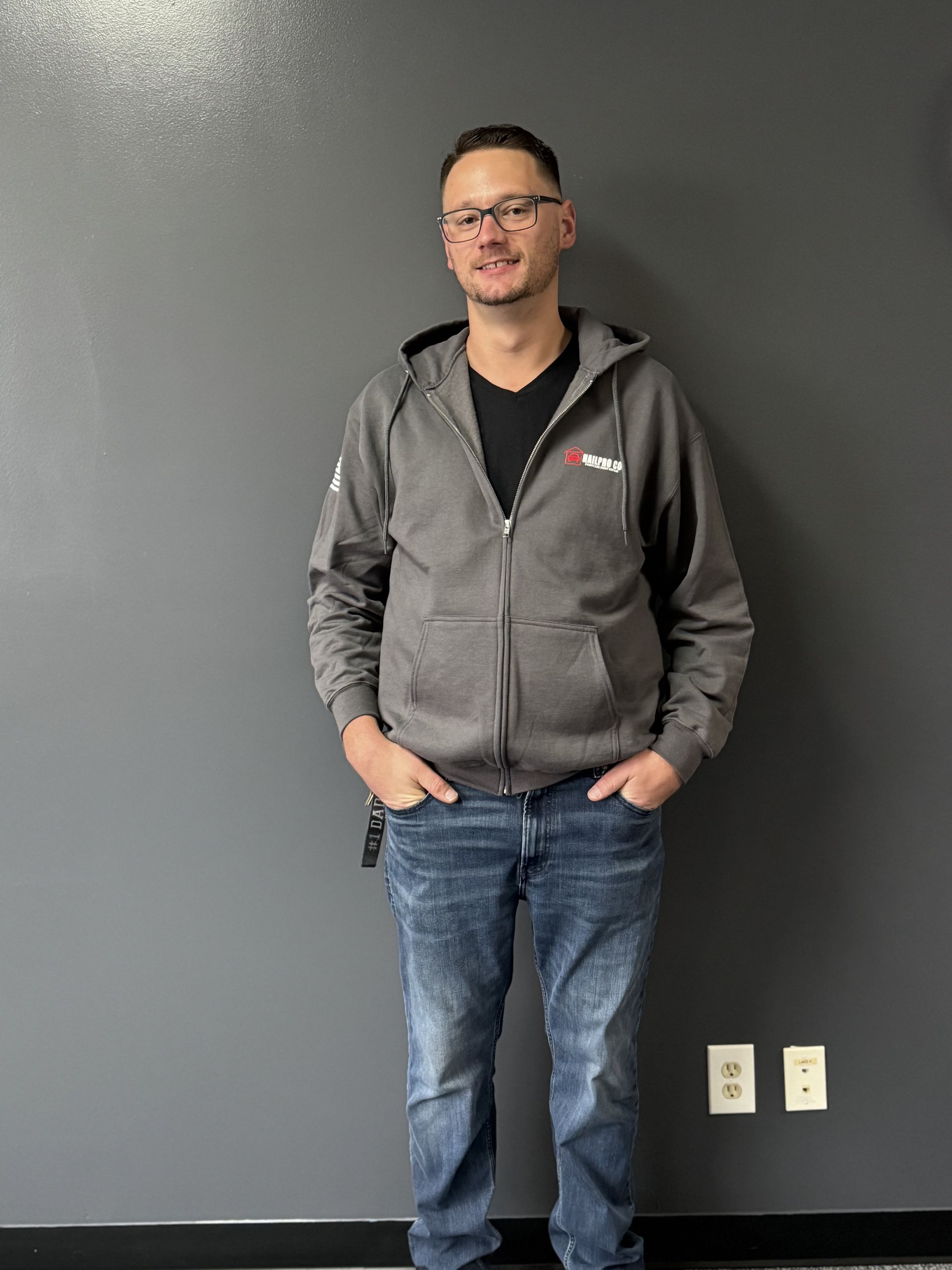 Man standing against a gray wall, wearing a gray hoodie and jeans, hands in pockets.