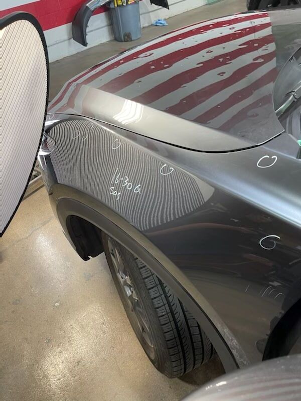 Black car fender with white sanding marks in a body shop, tire visible beside the wheel arch