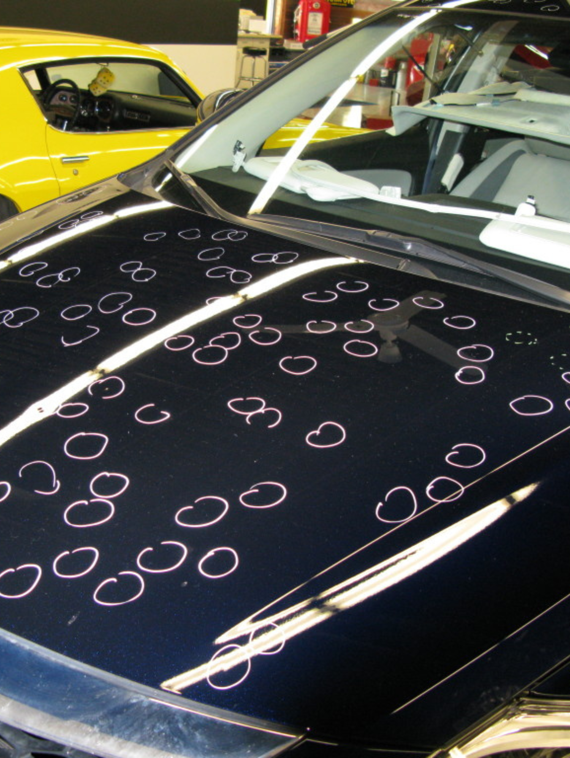 Black car hood marked with many pink chalk circles in a workshop, with a yellow car in the background