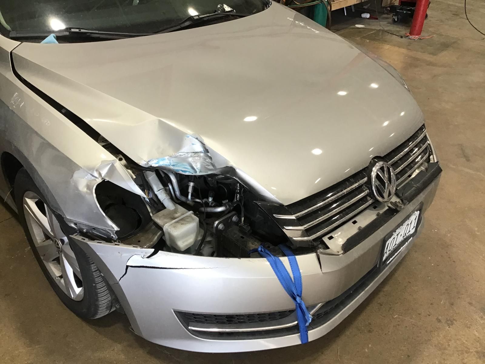 Front-left damage on a silver Volkswagen sedan, with a crumpled hood and smashed headlight in a garage.