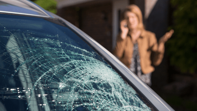 Cracked car windshield with spiderweb damage; blurred person standing in the background