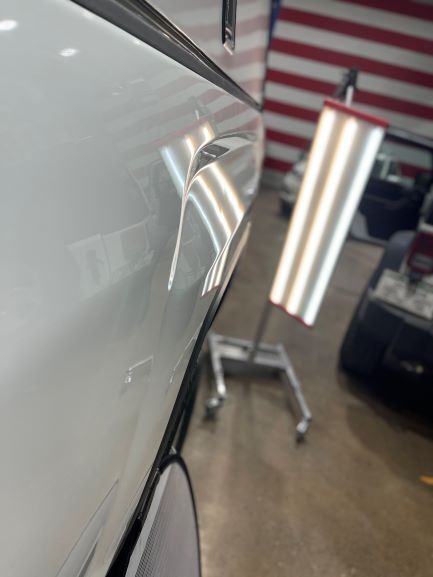 Close-up of a white car door with a dent, in a garage with bright lights and a tool cart.