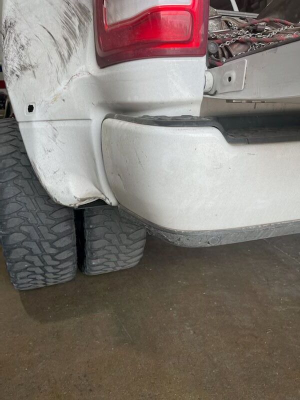 Close-up of a white pickup truck’s rear bumper and tire on a wet surface, with visible dirt and damage.