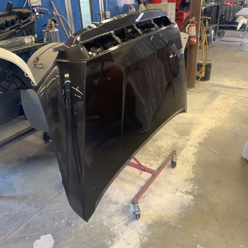 Black car hood leaning upright in an auto body shop, with a stand and tools nearby.