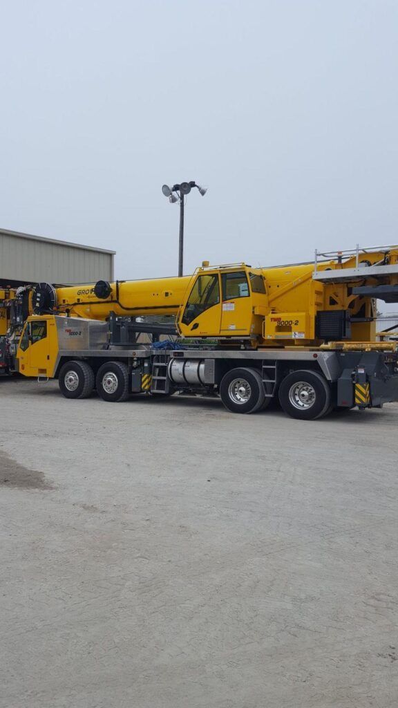 Yellow mobile crane truck parked on gravel lot near a warehouse under an overcast sky