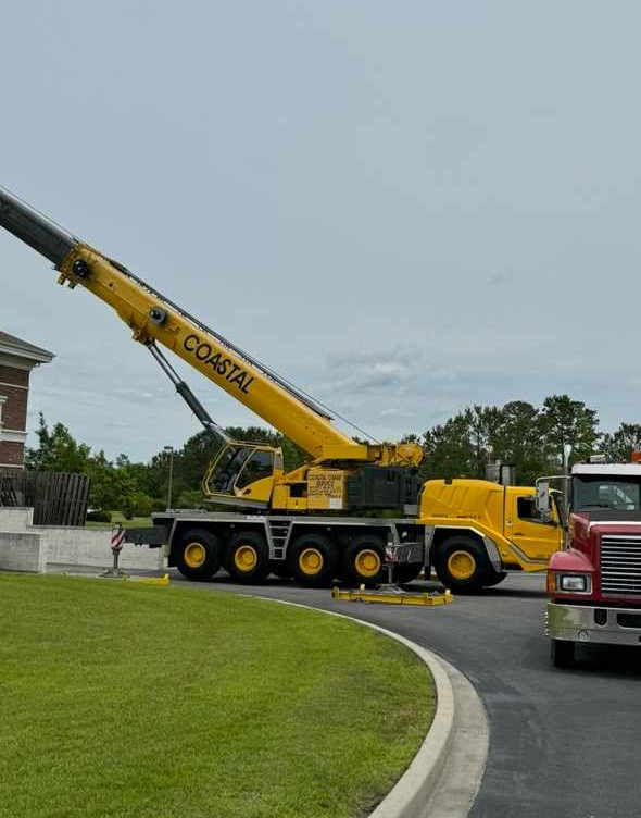 Yellow crane truck lifting boom on a driveway beside a lawn, with a red truck nearby under cloudy skies