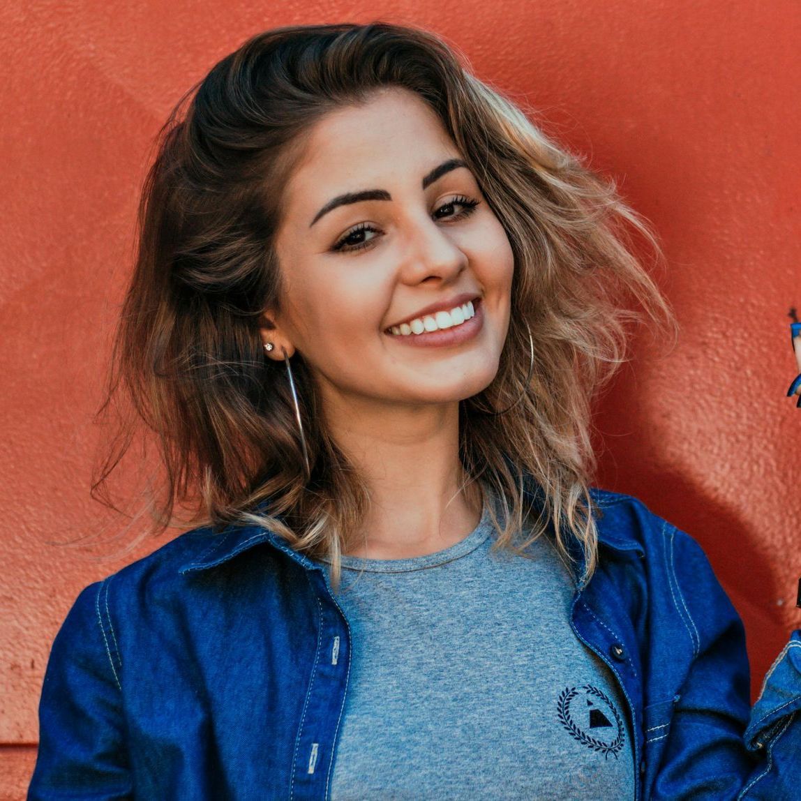 Woman smiling at the camera, wearing a blue denim jacket over a gray shirt, standing in front of an orange wall.