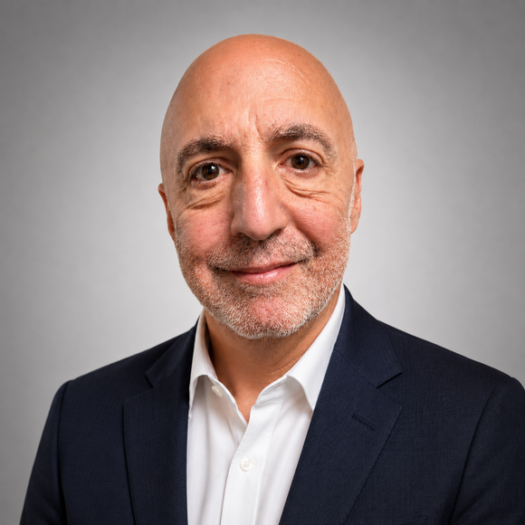 Portrait of a bald man in a dark blazer and white shirt, smiling against a gray background