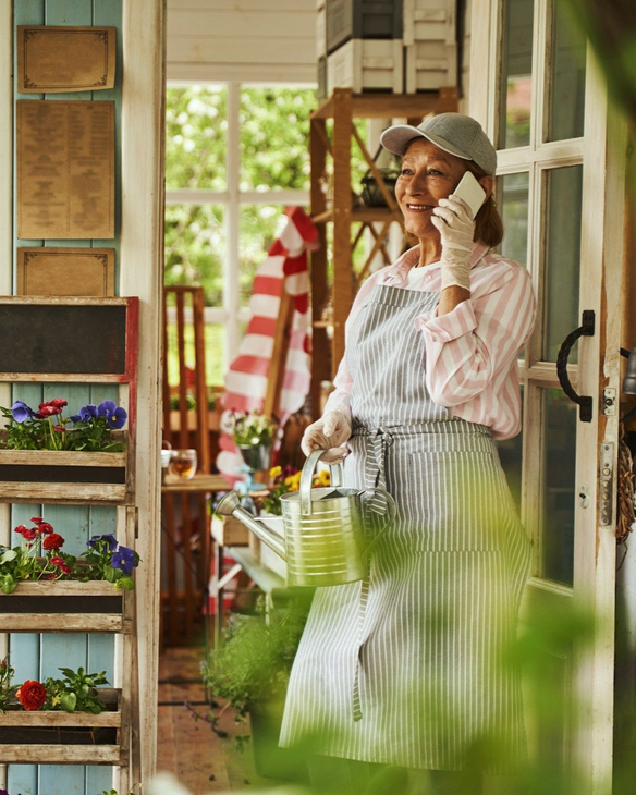 Woman in apron and cap standing in a porch doorway, talking on a phone beside flowers and a red-striped flag.