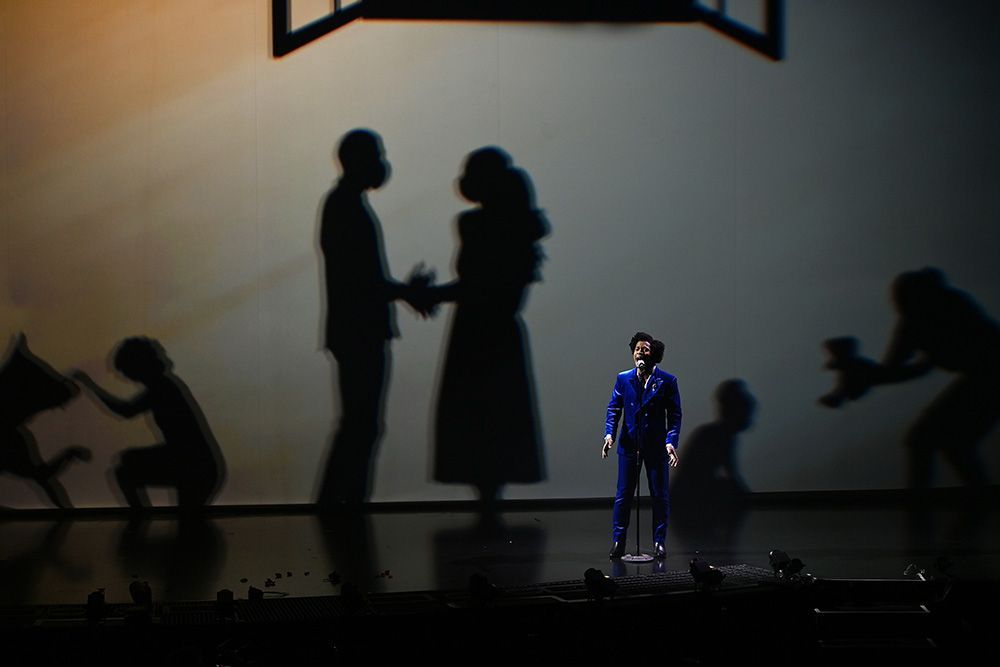 A lone person in a blue suit stands on a dark stage before shadowy figures in a tense scene.