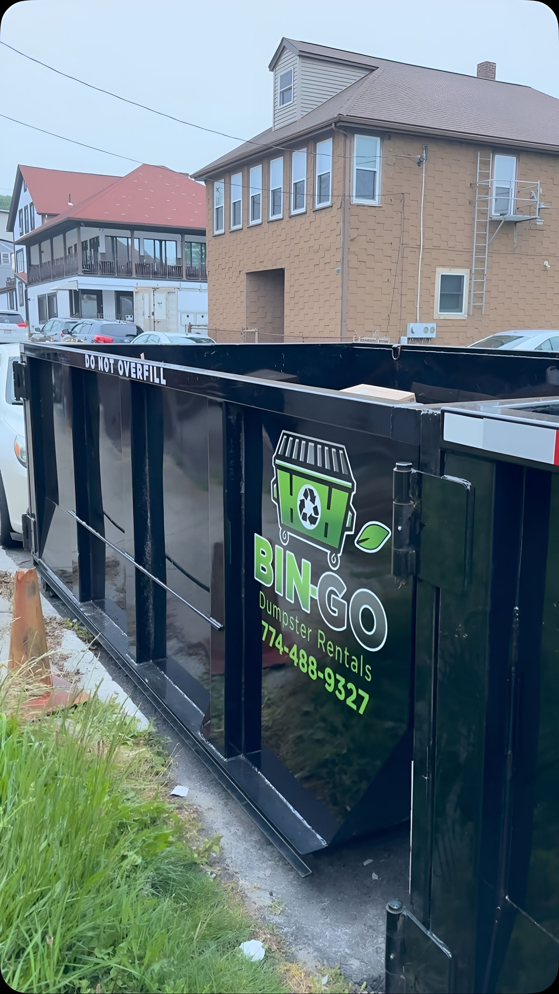 Black dumpster with green recycling sticker beside brick houses and a grassy curbside area.