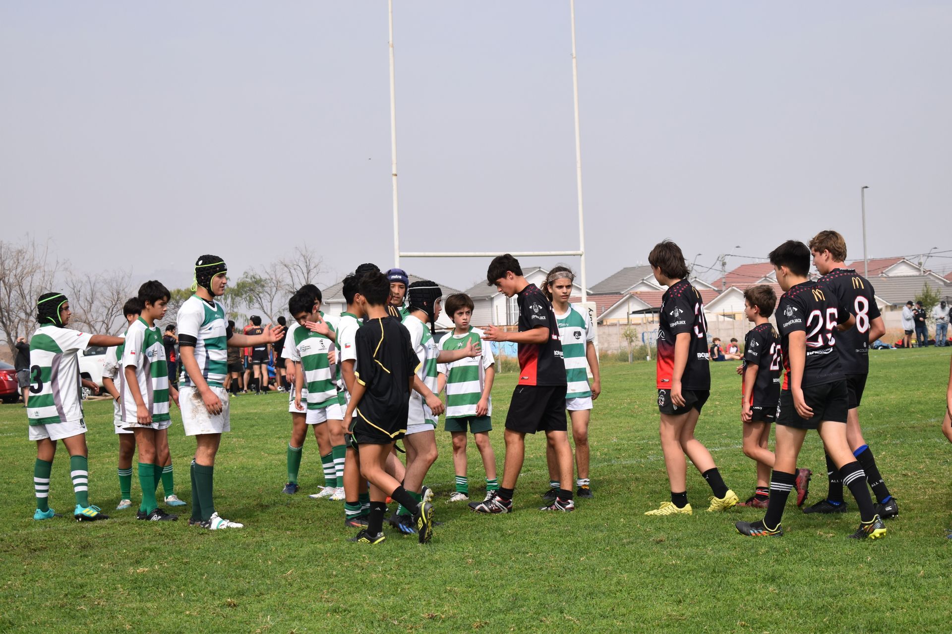 Los jugadores de rugby juveniles vestidos con camisetas verde/blancas y negras/naranjas se enfrentan en un campo de césped, cerca de los postes de la portería.