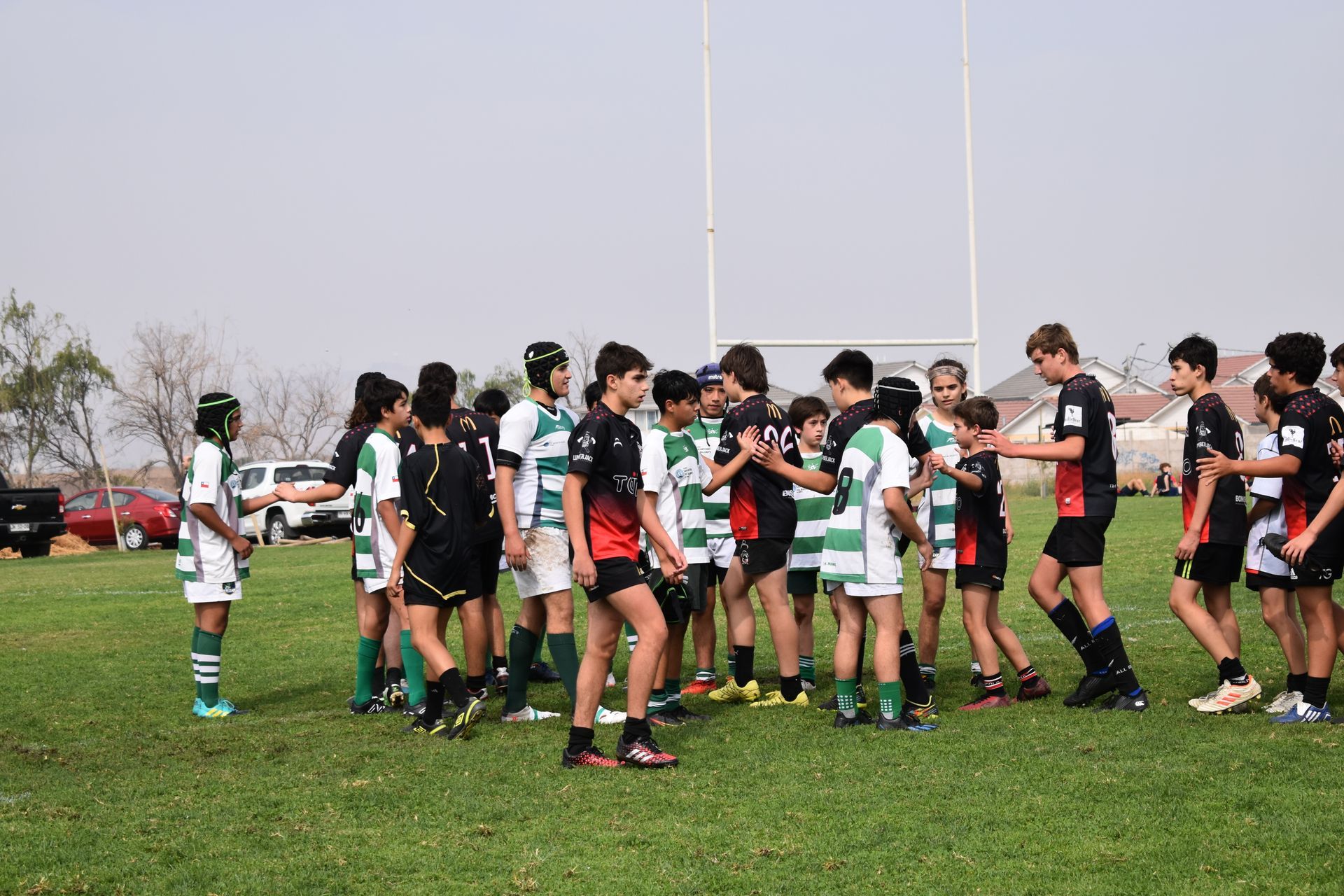Jugadores de rugby con uniformes verdes y negros se dan la mano en un campo de césped, con postes de portería al fondo.