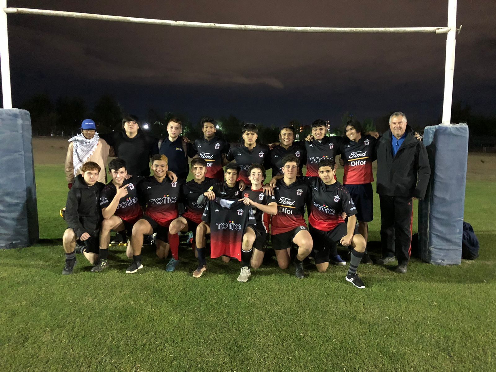 Un equipo de rugby posa en un campo de noche. Jugadores con uniformes negros y rojos se paran frente a una portería.