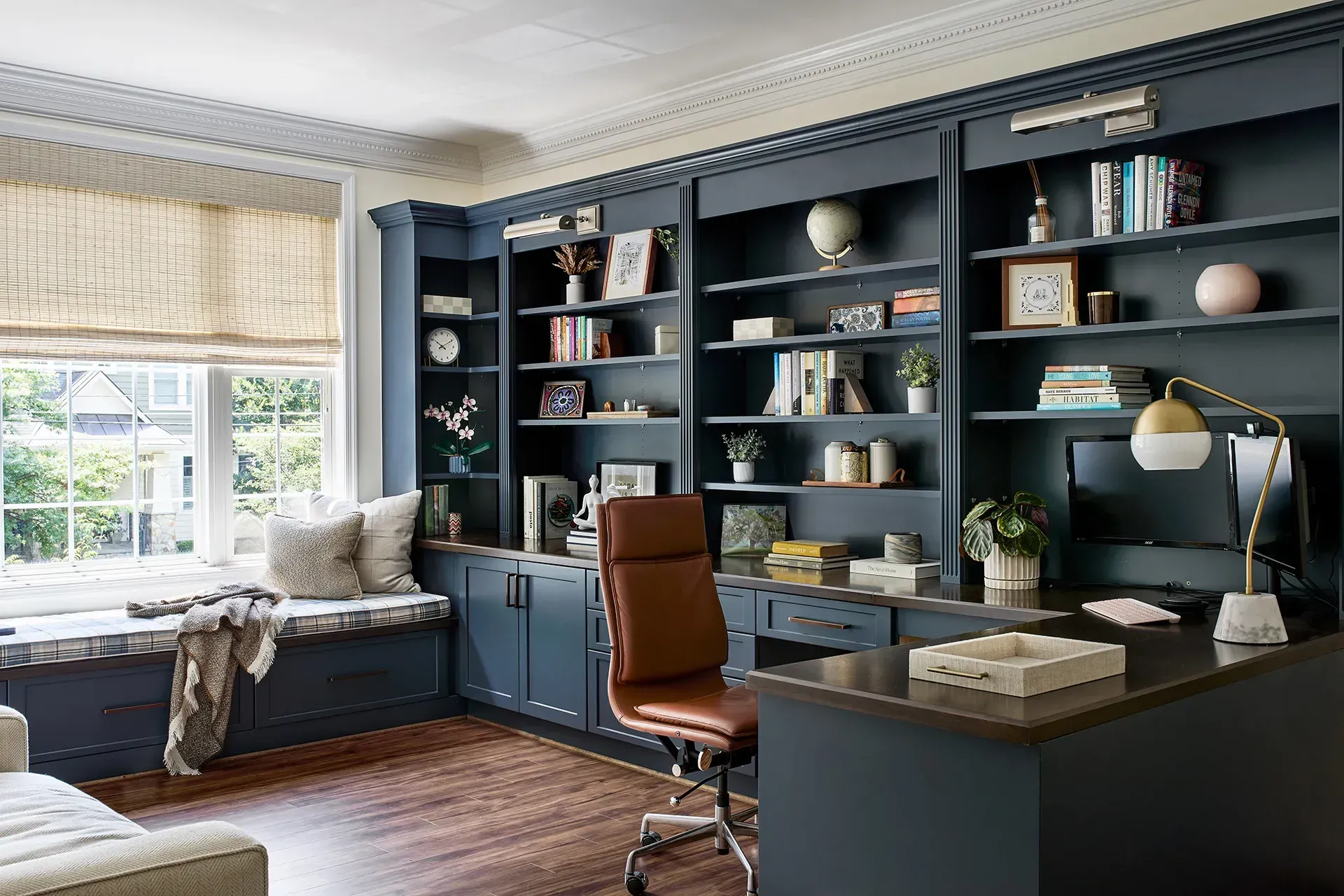 Home office with navy blue built-in bookshelves and desk, brown leather chair, and window seat.