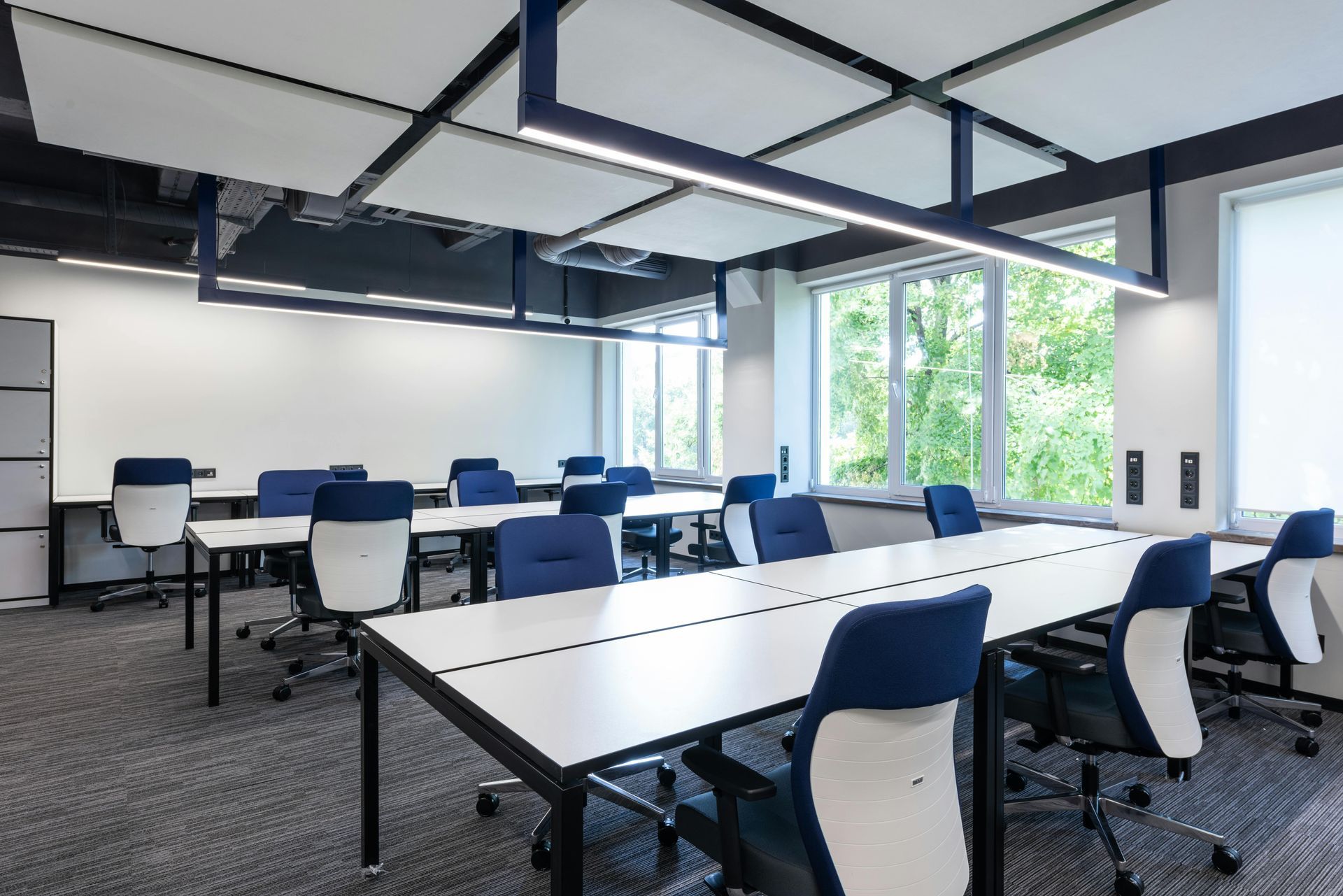 Office with long white desks, blue chairs, and sound-dampening ceiling tiles. Natural light from large windows.