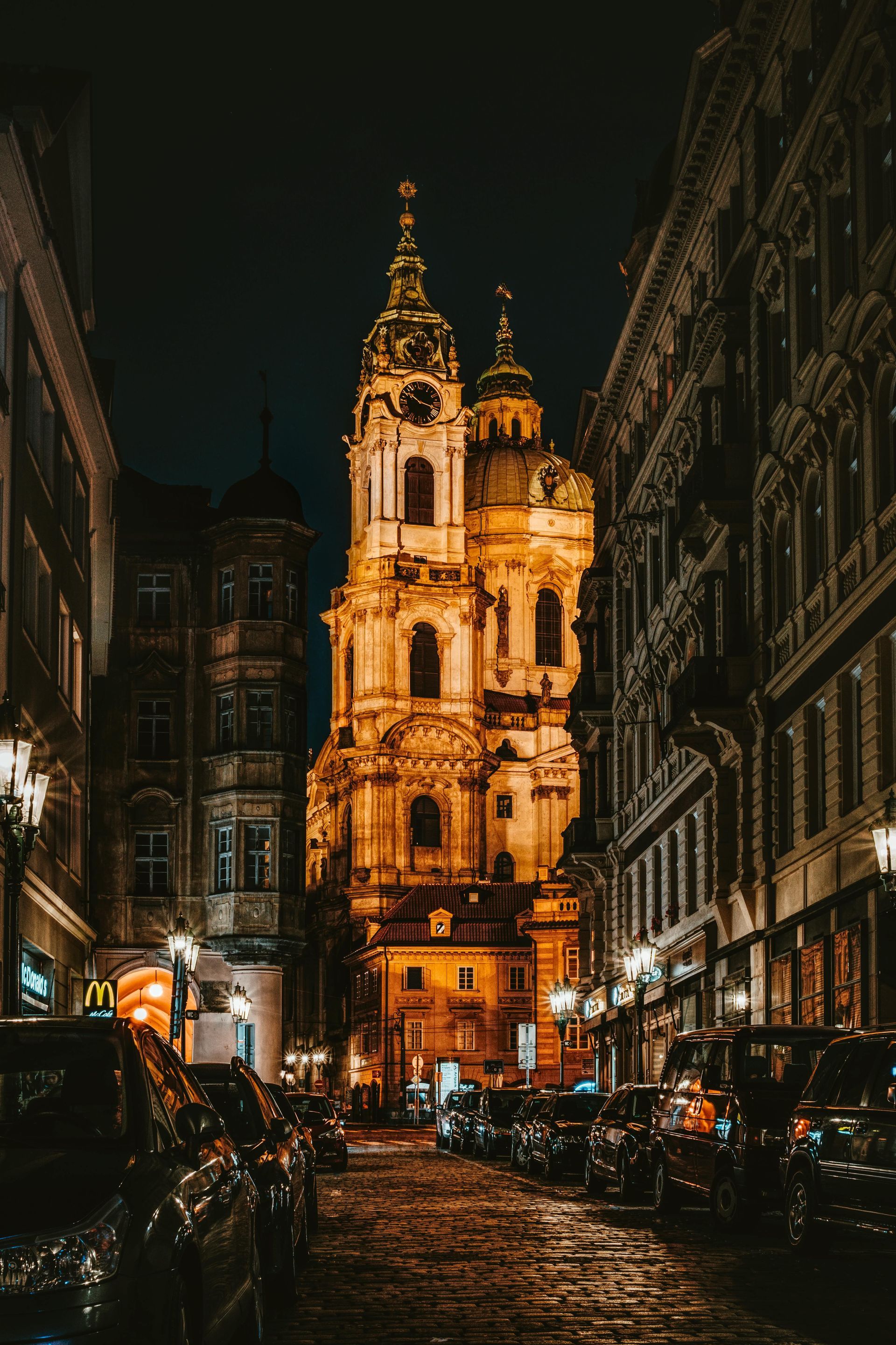 Church at night, illuminated, between buildings on a cobblestone street.