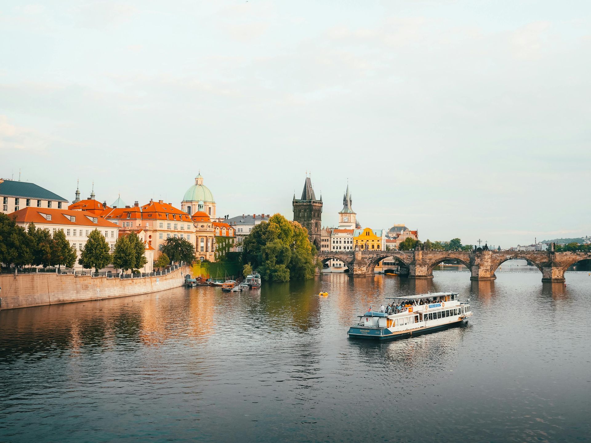 Prague cityscape with Vltava River, Charles Bridge, and tour boat under a light sky.