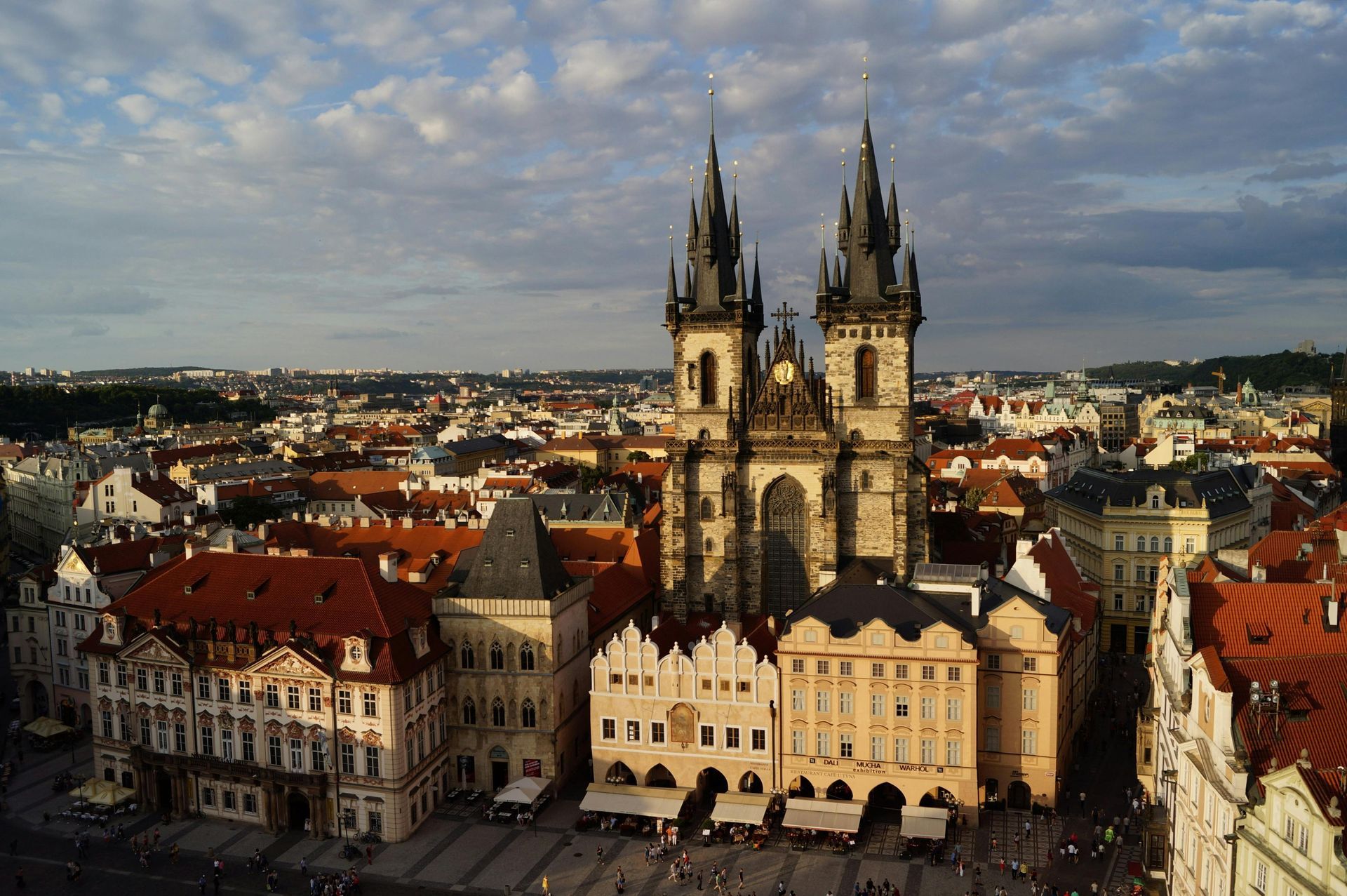 Church of Our Lady Before Týn, Prague, with twin Gothic towers. Buildings with orange roofs surround the church under a cloudy sky.