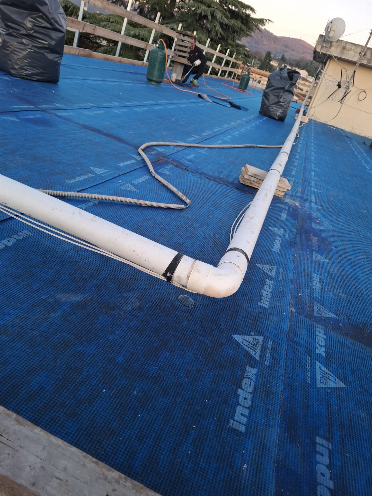 Area di lavoro sul tetto blu con tubi in PVC bianchi, corde e alcune persone in lontananza al tramonto.