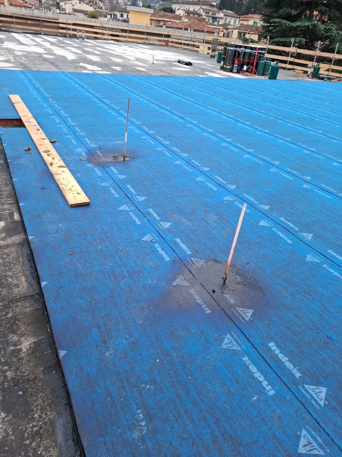 Tetto blu impermeabilizzato in costruzione, con assi di legno, tubi e gli edifici circostanti sullo sfondo.