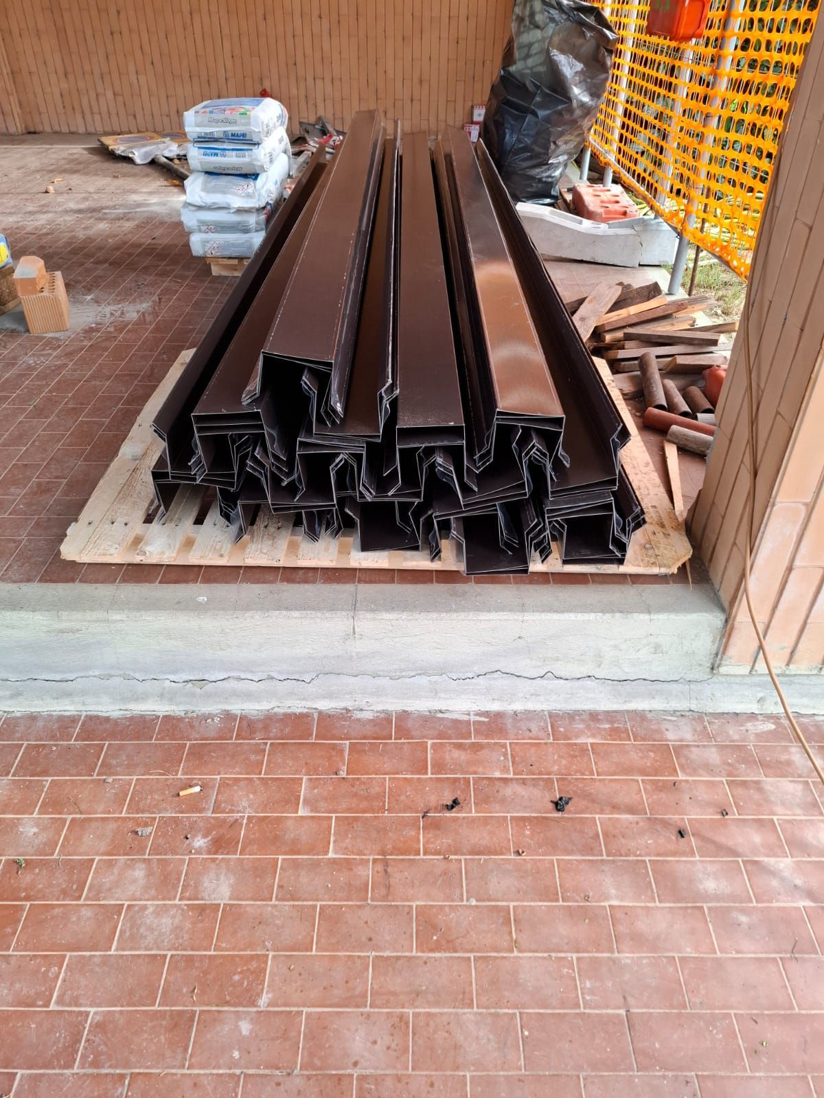 Travi metalliche scure impilate su un pallet in un magazzino o in un cantiere, con attrezzi e materiali nelle vicinanze.
